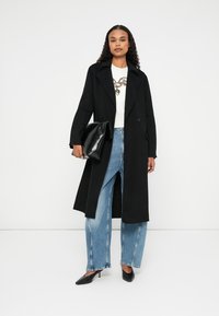 Abrigo negro de corte largo con grandes solapas, botones de doble botonadura, combinado con una camiseta blanca, jeans azules de corte amplio y tacones negros puntiagudos.