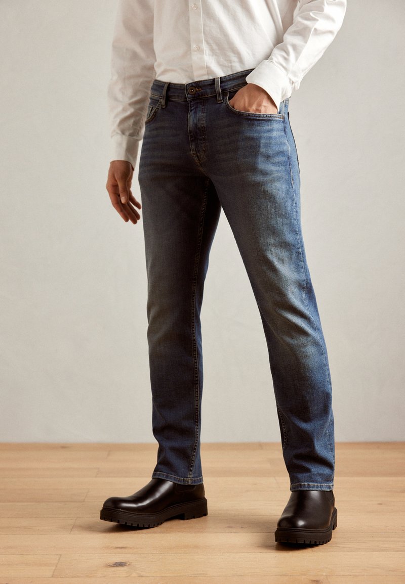 Blue denim jeans with a straight-leg cut, featuring front pockets, faint fading, and a classic five-pocket design, paired with black shoes.