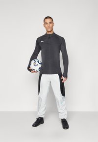 Schwarzes, geripptes, langärmliges Sporttop mit hohem Kragen, kombiniert mit weißen Hosen mit schwarzen seitlichen Akzenten. Das Modell hält einen Fußball.