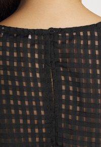 Tessuto nero con texture e motivo a griglia, dotato di chiusura a bottone sul retro e di una piccola fessura all'escursione del collo.