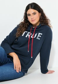 Marineblauwe hoodie met zilveren "TRUE" tekst, rode koordjes, voorzak en een relaxte pasvorm. Draagt bij blauwe jeans, weergegeven tegen een witte achtergrond.