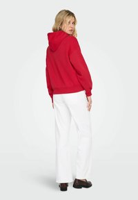 Femme en sweat à capuche rouge et pantalon blanc debout, regardant par-dessus son épaule, portant des mocassins marron foncé sur un fond uni.