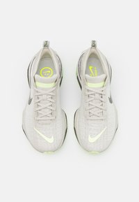 Gaiši pelēki sporta apavi ar teksturētu virsu, atstarojošiem akcentiem, zaļiem zoliem un izteiksmīgu baltu Nike simbolu.