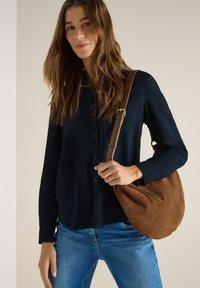 Femme portant une chemise bleu foncé boutonnée et un jean bleu, tenant un sac à bandoulière en daim marron avec une sangle en cuir.