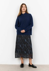 Maglione zip-up navy con colletto alto, abbinato a una gonna nera con motivi astratti e pieghe, completato da mocassini neri.