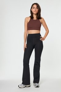 Ženska stoji z eno roko na boku, oblečena v rjav cropped top, črne visokotlačne flare legice in srebrne športne čevlje na beli podlagi.
