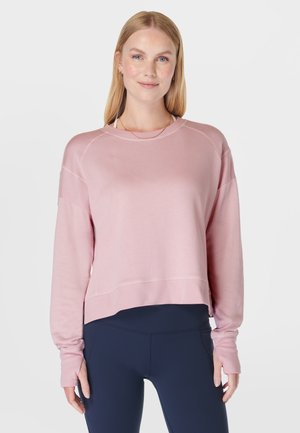 Vrouw met stijl blond haar, gekleed in een roze sweatshirt met lange mouwen en donkeblauwe leggings, staand tegen een effen lichte achtergrond.