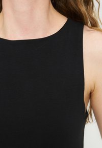 Κοντινό πλάνο ενός ατόμου που φορά ένα μαύρο αμάνικο τοπ με ψηλή, ίσια γραμμή στο λαιμό, που δείχνει τον λαιμό και τους ώμους.