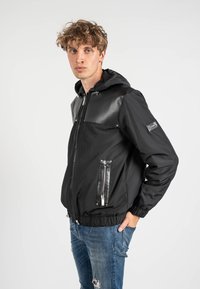 Giacca bomber nera con pannello frontale lucido, cappuccio e due tasche con zip. Presenta polsini e orlo a costine, realizzata in tessuto morbido.
