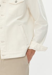 Jachetă din denim alb cu două buzunare pe piept, cusături vizibile și un design de mansetă pliabil. Fabrică texturată fără cusături, cu un finisaj ușor.