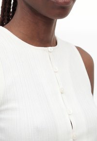 Gros plan d'une personne à la peau foncée portant un haut blanc côtelé sans manches avec cinq boutons blancs sur le devant.