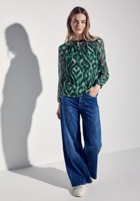 Grüne gemusterte Bluse mit langen Ärmeln, kombiniert mit weiten blauen Jeans und weißen Sneakern. Der Schatten der Figur ist an der Wand sichtbar.
