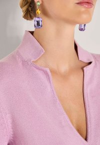 Nahaufnahme einer Person, die ein lilafarbens Strickoberteil mit einem Halsausschnitt und auffälligen Ohrringen mit grünen, gelben und lila Edelsteinen trägt.
