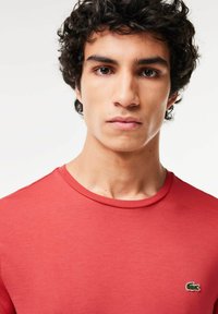 Röd t-shirt med rund halsringning och korta ärmar. Har en liten grön krokodil-logotyp på vänster bröst. Slät tygtextur.