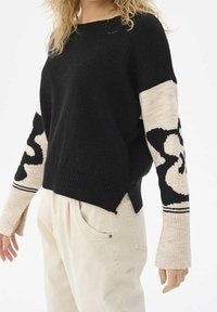 Pull en maille noire avec des manches beiges contrastantes ornées d'un motif noir audacieux. Il présente une coupe décontractée et des fentes latérales à l'ourlet.