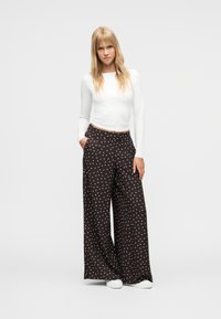 Νεαρή γυναίκα με μακριά ξανθά μαλλιά, φοράει λευκό crop top με μακριά μανίκια και μαύρο παντελόνι με άνετη φαρδιά γραμμή και λευκές πουά, στεκόμενη.