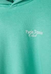 Sudadera verde menta con una textura suave y capucha con cordón. Presenta texto impreso: "Club de Tenis de París" en blanco. Diseño sencillo.
