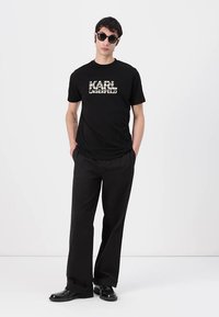 Juodas medvilninis marškinėliai su dideliu logotipu "KARL LAGERFELD" kontrastingame rašte, derinami su laisvais juodais kelnių ir blizgiais bateliais.