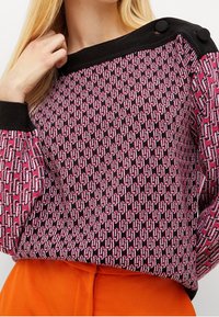 Gebreide trui met een geometrisch roze en zwart patroon, met een zwarte ribbelkraag en knopenaccenten op de schouder.