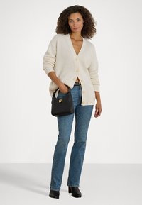 Cardigan de malha de cor creme com decote em V e corte oversized. Apresenta grandes botões, combinado com jeans azul, uma bolsa preta e botas de tornozelo.