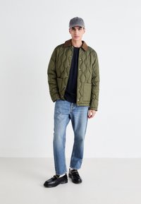 Quilted olive green jacket with brown collar, two front pockets, worn over a navy shirt, paired with light blue jeans and black shoes.