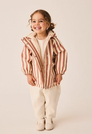 Lächelndes kleines Kind mit lockigem Haar in einer gestreiften rosa-beigen Kapuzenjacke, cremefarbenen Hosen und beigen Sneakers, stehend vor hellem Hintergrund.