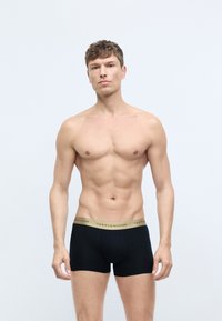 Sorte boxer shorts med en guld "TOMMY HILFIGER" logo-elastik, der har en blød stofstruktur og en tætsiddende pasform. Model står med en neutral ansigtsudtryk.