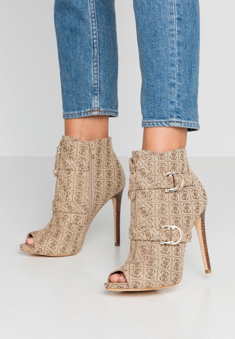 Beige nilonit korku ankle botlar, tekrar eden logo deseniyle dokulu bir kumaşa sahip, açık burunlu ve yan tarafında çift tokalı detaylar bulunuyor.
