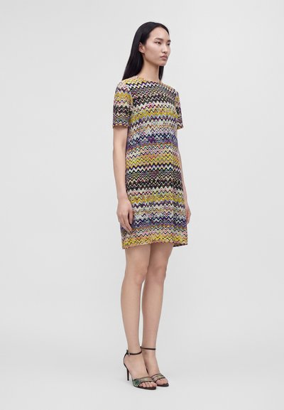 Femme debout portant une robe à manches courtes à motif zigzag multicolore avec des sandales ouvertes à talons hauts, sur un fond uni.