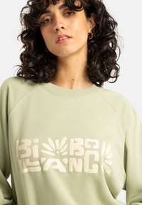 Groene sweatshirt met een ronde hals en raglanmouwen. Voorzien van een groot, verhoogd, crèmekleurig logo ontwerp aan de voorkant.