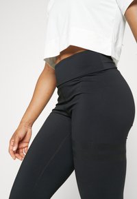 Schwarze hochgeschnittene Leggings aus elastischem Material, mit Netzdetails und genähten Akzenten an den Seiten.