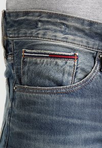 Jeans i denim med en sliten blå finish, med en liten bakficka med röd, vit och blå accentrand och kopparnitar.