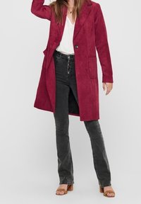 Abrigo largo de pana burdeos con cuello de muesca, cierre de un solo botón, bolsillos laterales, llevado sobre una blusa blanca y jeans negros acampanados.