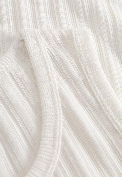 Hvitt ribbestrikket stoff med teksturerte striper og en glatt, avrundet halslinning. Materialet virker mykt og lett, ideelt for lag-på-lag.