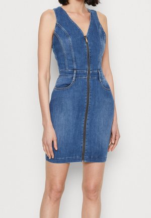 Robe en jean bleue sans manches avec fermeture éclair à l'avant, taille cintrée et poches, portée par une personne debout aux cheveux courts et foncés.