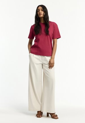 Frau mit langen dunklen Haaren, die ein rotes kurzärmeliges Hemd, weite beige Hosen und braune Sandalen mit Absatz trägt, steht mit einer Hand in der Tasche.