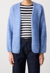 Cardigan bleu clair, à ouverture devant, en matériau doux et texturé avec de longues manches légèrement bouffantes. Porté par-dessus une chemise rayée et un jeans foncé.