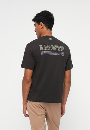 Juodas trumpomis rankovėmis Tmaras su didelėmis daugiaspalvėmis raidėmis, užrašytomis ant nugaros "LACOSTE". Virš apykaklės yra mažas logotipas.