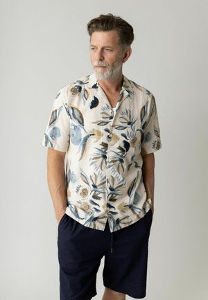 Mand med gråt skæg iført kortærmet, knaplukket blomsterprintskjorte og mørke shorts, stående med hænderne i lommerne og kiggende mod venstre.