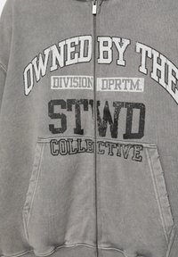 Szürke cipzáras kapucnis pulóver első zsebekkel, fehér és fekete, kopott hatású felirattal: "OWNED BY THE STWD COLLECTIVE" és kisebb betűkkel "DIVISION DPRTM."