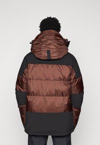 Giubbotto imbottito in nero e marrone con un design trapuntato, grande cappuccio e due tasche laterali. Presenta materiali lisci e testurizzati.