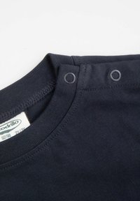 Zbliżenie na niebieską koszulkę dla niemowląt z podwójnym zapięciem na zatrzaski i widoczną metką marki wewnątrz kołnierzyka.