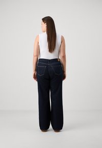 Jeans de pierna ancha de color azul oscuro, fabricados en denim, con una cintura alta, dos bolsillos traseros y detalles de costura visibles. Combinados con una blusa sin mangas blanca.