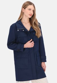 Marineblå lang frakke med opretstående krave, bryst- og sidelommer, ærmer med knapper og lynlåslukning. Glat tekstur og stof.