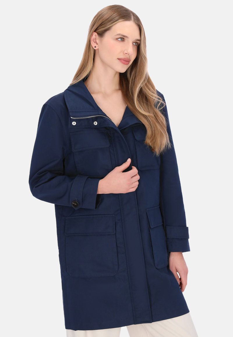 Marineblå lang frakke med opretstående krave, bryst- og sidelommer, ærmer med knapper og lynlåslukning. Glat tekstur og stof.