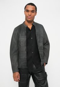 Chaqueta de piel sintética de color gris oscuro con cuello alzado, que presenta un cierre de cremallera y un sutil estampado de logo. Combinada con una camisa negra de botones.