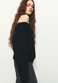 Schwarzer schulterfreier Pullover mit geripptem Ausschnitt, lockerer Passform und langen Ärmeln. Kombiniert mit dunkelgrauen Jeans. Glatte Textur.