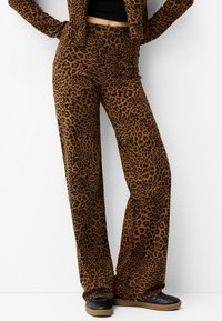 Hnědé kalhoty s leopardím potiskem, vyrobené z měkké tkaniny, s rozšířeným střihem a elastickým pasem, doplněné černými botami.
