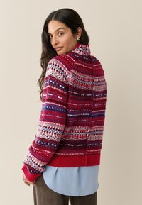 Gebreide rode trui met witte, blauwe en paarse patronen; hoge kraag, lange mouwen, geribbelde manchetten en zoom; gelaagd over een blauw shirt.