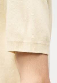 Tissu tricoté crème à manches courtes avec motif texturé, bords de poignets ajustés et peau visible de l'avant-bras sous la manche.